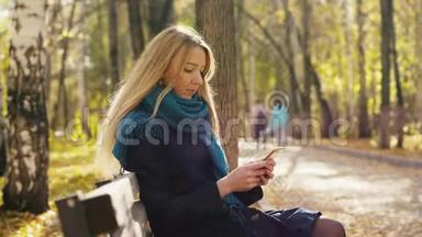 年轻漂亮的金发女人<strong>坐在</strong>长凳上，在晴天的秋日<strong>公园里</strong>使用智能手机。 3840x2160